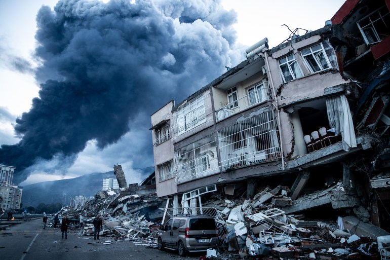 HATAY, TURKEY - FEBRUARY 07: Smoke billows from Iskenderun Port fire as people walk past collapsed buildings on February 07, 2023 in Iskenderun, Turkey. A 7.8-magnitude earthquake hit near Gaziantep, Turkey, in the early hours of Monday, followed by another 7.5-magnitude tremor just after midday. The quakes caused widespread destruction in southern Turkey and northern Syria and were felt in nearby countries. (Photo by Burak Kara/Getty Images)