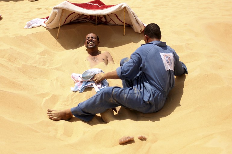 A patient grimaces as a worker covers his body in hot sand in Siwa, Egypt, August 11, 2015. In the searing heat of summer in western Egypt, at the hottest time of the day, sufferers of rheumatism, joint pain, infertility or impotence lie buried neck-deep in the sand of Siwa near Dakrour Mountain. Locals say taking a sand bath is a natural therapy with powers to cure many medical conditions. Patients relax in the shade before treatment, which includes massages by the feet of health workers after they submerge their patients up to their neck in the desert. Patients drink mint tea in tents following the treatment. REUTERS/Asmaa Waguih PICTURE 8 OF 24 FOR WIDER IMAGE STORY "THE HOT SAND BATHS OF SIWA". SEARCH "ASMAA SIWA" FOR ALL IMAGES FOR BEST QUALITY IMAGE ALSO SEE: GF20000090219