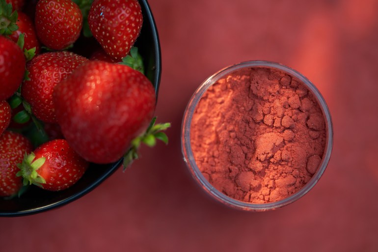 Opened container of 80 gram of freeze-dried strawberry powder. Bowl of strawberries. Red surface. Light effect. High point of view.