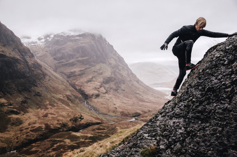 A free runner climbs a steep mountain rock face with the view of the three sisters mountains in the background.