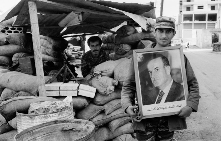 Portrait of an unidentified Syrian soldier as he poses, with a framed portrait (of President Hafez al-Assad), beside the sandbags of a covered checkpoint on an unidentified street, Beirut, Lebanon, June 1987. Visible behind him is a fellow soldier in the shelter with a machine gun. (Photo by Tom Stoddart/Getty Images)