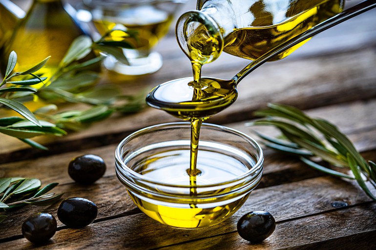 Pouring extra virgin olive oil from a spoon to a glass container. Olive branches comes from the left and right. Some olive oil bottles are out of focus at background. Black olives are on the table and complete the composition. The composition is on a rustic wooden kitchen table. Predominant colors are gold, green and brown. High resolution 42Mp studio digital capture taken with Sony A7rII and Sony FE 90mm f2.8 macro G OSS lens