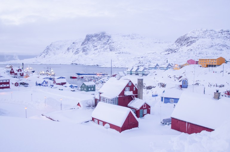 Panorama from top of hill on arctic settlement town in Greenland during freezing dark day in the middle of nowhere buried in snow with a view of colorful houses and frozen bay and mountains behind