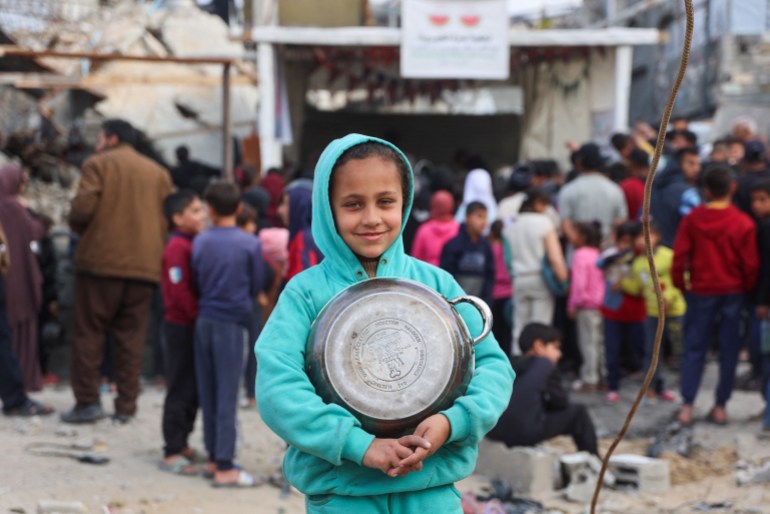 A Palestinian girl poses for a picture as she heads to queue at a charity kitchen to receive a cooked food portion in Beit Lahia in the northern Gaza Strip, ahead of the iftar fast-breaking meal during the Muslim holy month of Ramadan on March 9, 2025. Israel's energy minister said on March 9 he had given instructions to stop supplying electricity to Gaza, a week after Israel blocked all aid into the war-ravaged Palestinian territory. (Photo by Omar AL-QATTAA / AFP)